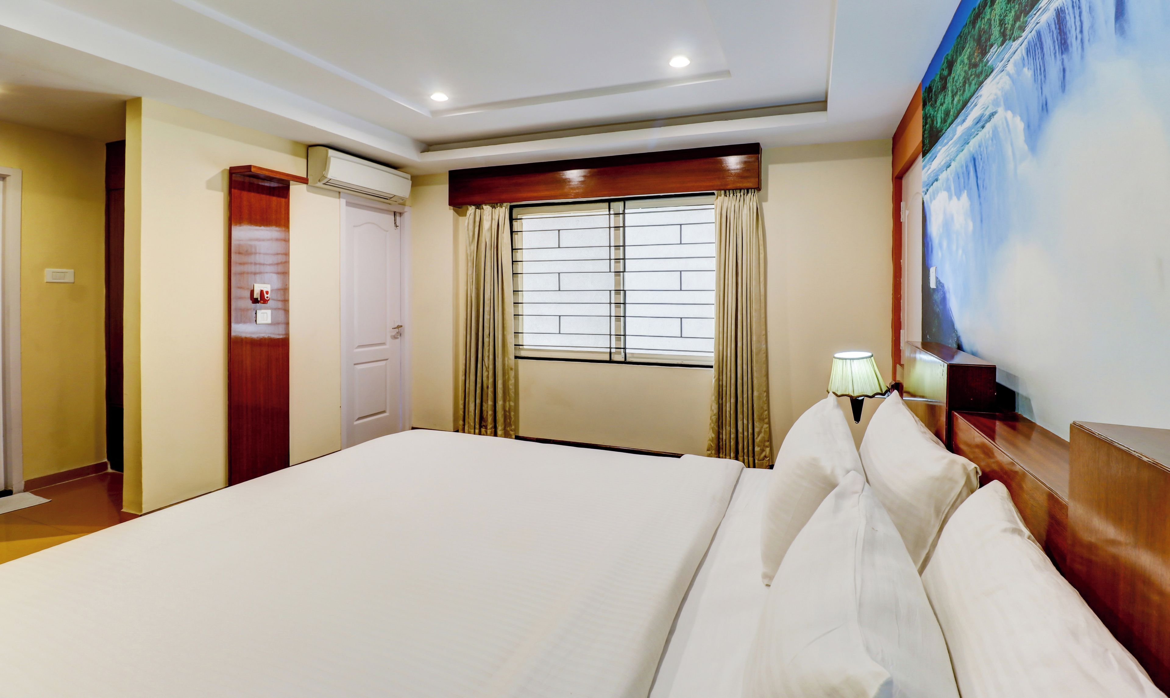 A hotel room with a large bed, a window with curtains, an air conditioning unit, and a wall-mounted lamp.
