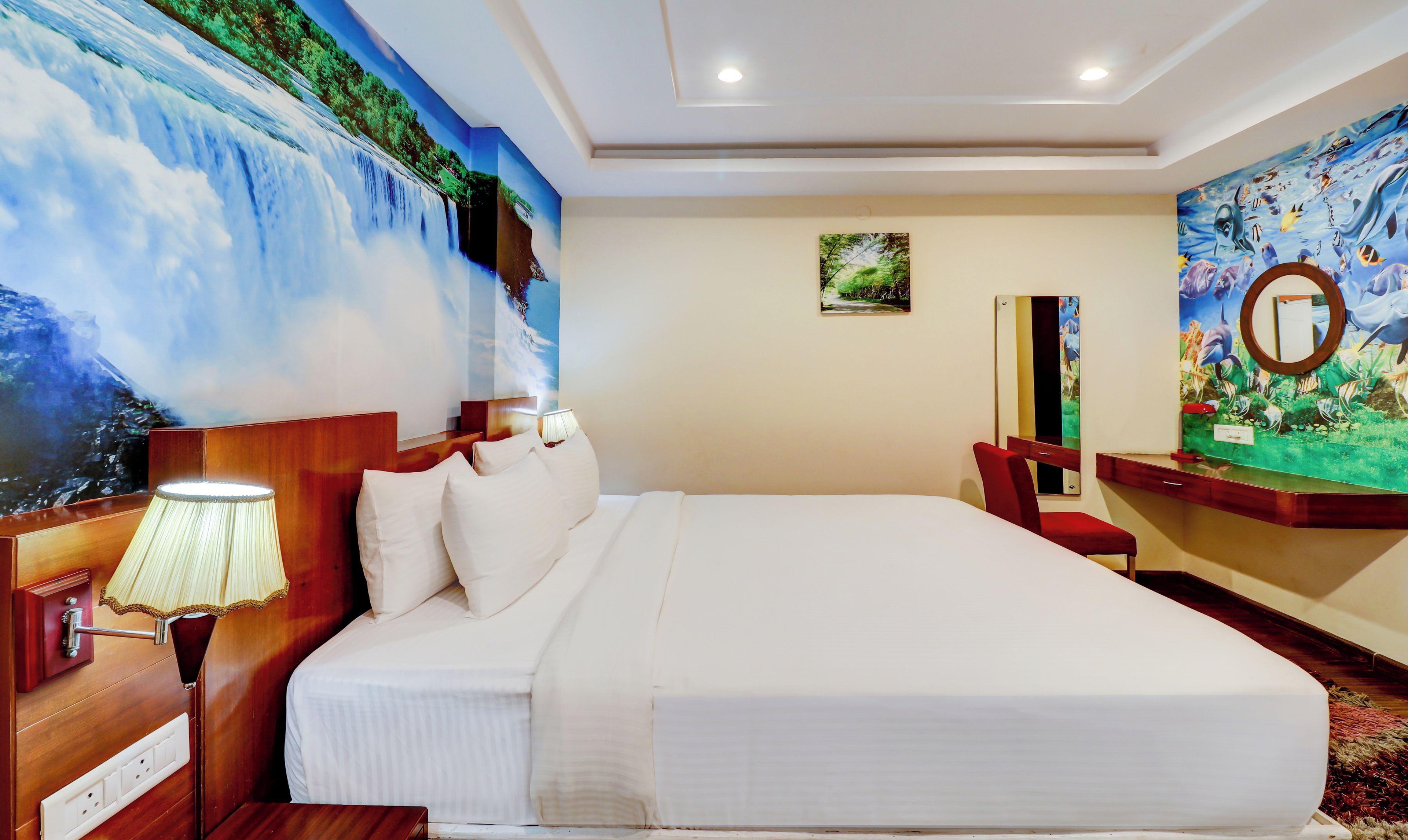 Una habitación de hotel con una cama grande, una mesita de noche con lámpara, un escritorio con espejo y un mural en la pared que representa una cascada.