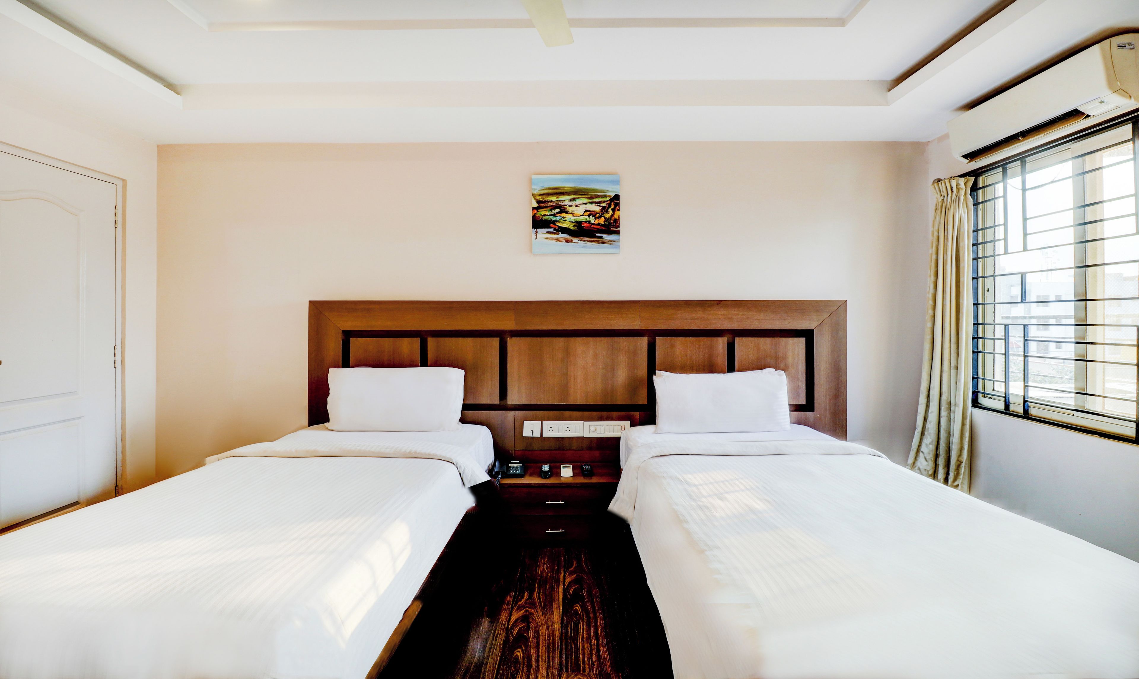 Habitación de hotel con dos camas, cabecera de madera, un cuadro en la pared y una ventana con cortinas.