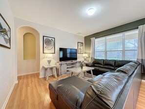 A living room with a leather sofa, a TV, a small table with a lamp, and artwork on the walls. - PROMO Magical 6 BR Villa, Pool/Spa, Windsor Hills - by Blue Sky Villas (Kissimmee)