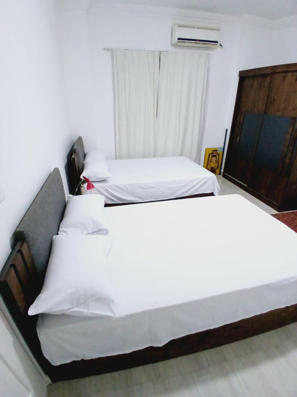Habitación con dos camas, ropa de cama blanca, cabecero de madera, aire acondicionado y una ventana con cortinas.