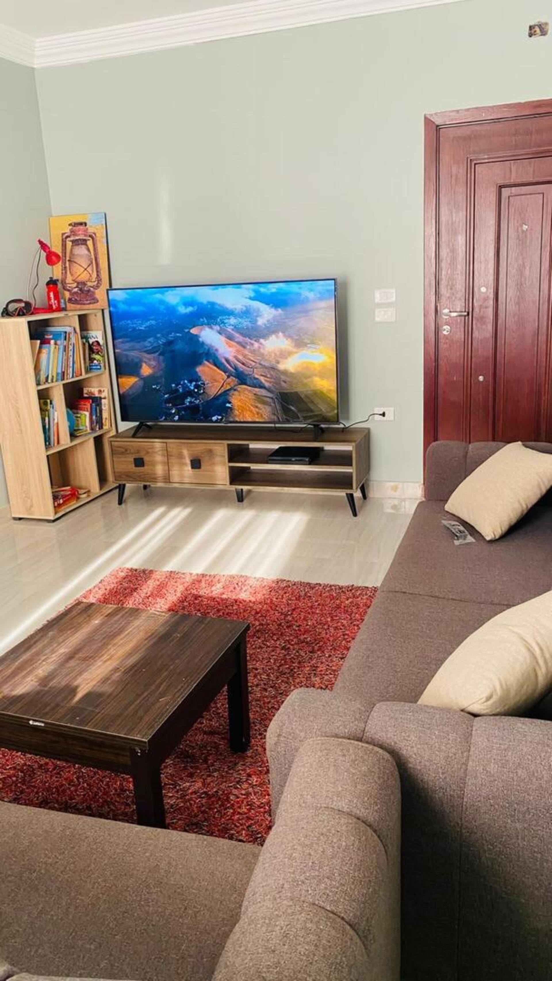 A living room with a flat-screen TV on a wooden stand, a bookshelf, a red rug, a brown sofa, and a wooden door.