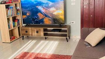 Een woonkamer met een flatscreen televisie op een houten standaard, een boekenkast, een rood vloerkleed, een bruine bank en een houten deur.