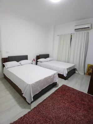 A hotel room with two single beds, a red rug, white curtains, and an air conditioning unit. - Modern Apartment at Hurghada City Palace (Hurghada)