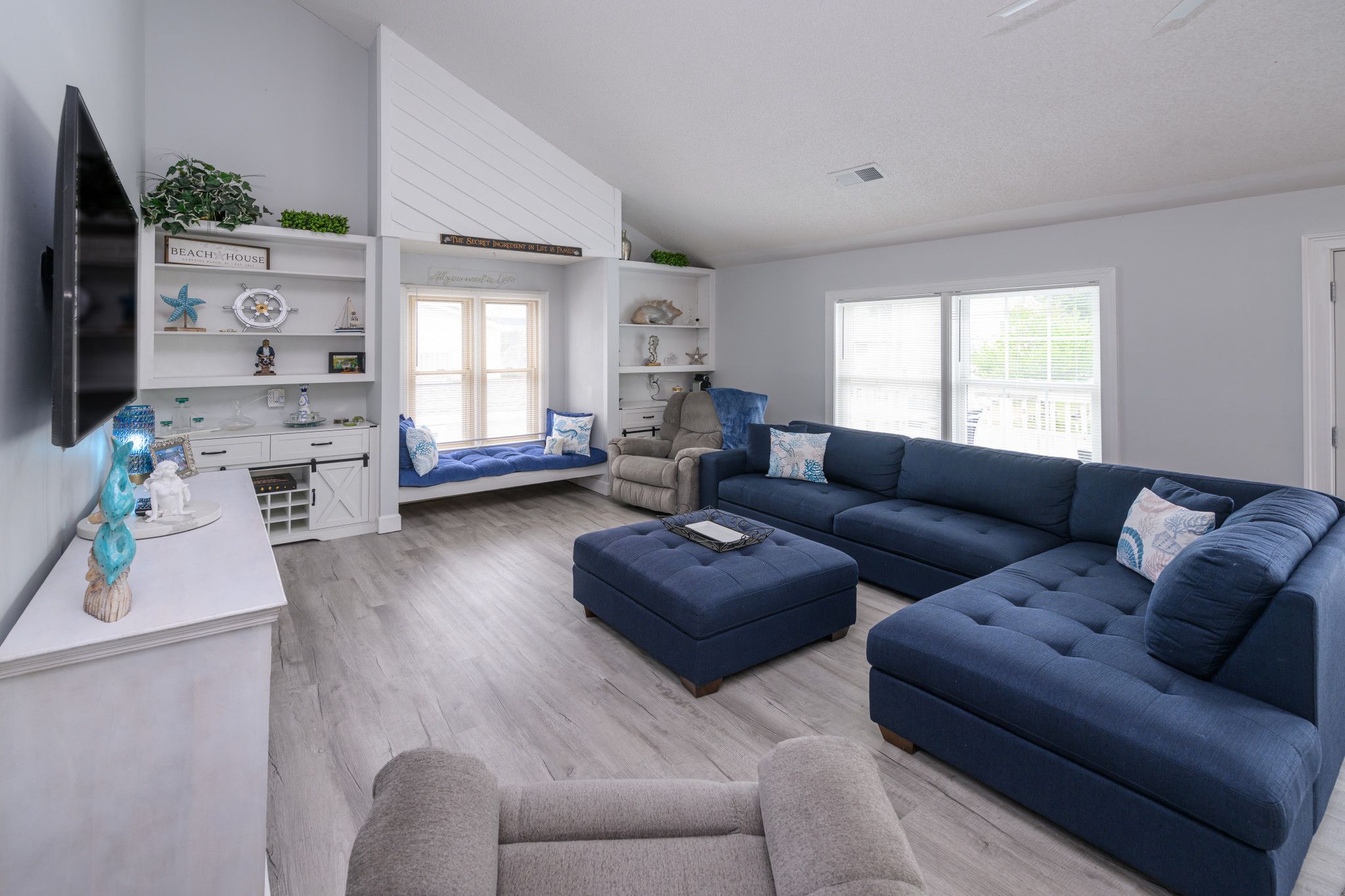 A modern living room with a sectional sofa, a small armchair, a coffee table, and a TV mounted on the wall. There are built-in shelves with decorative items and a window with blinds.
