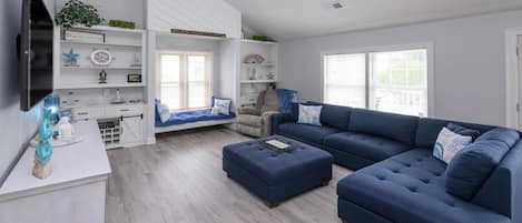 A modern living room with a sectional sofa, a small armchair, a coffee table, and a TV mounted on the wall. There are built-in shelves with decorative items and a window with blinds.