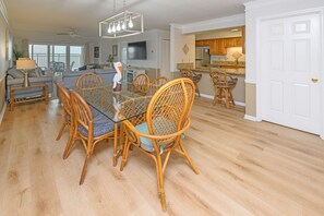 A spacious living area with a dining table, wicker chairs, and a modern kitchen in the background. - 2BR Oceanfront Condo with Elevator and Shared Pool! South Shores II 203 (Surfside Beach)