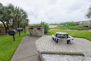 An outdoor patio with a stone barbecue, picnic tables, and a small brick structure. - 2BR Oceanfront Condo with Elevator and Shared Pool! Surfwatch II 302 (Surfside Beach)