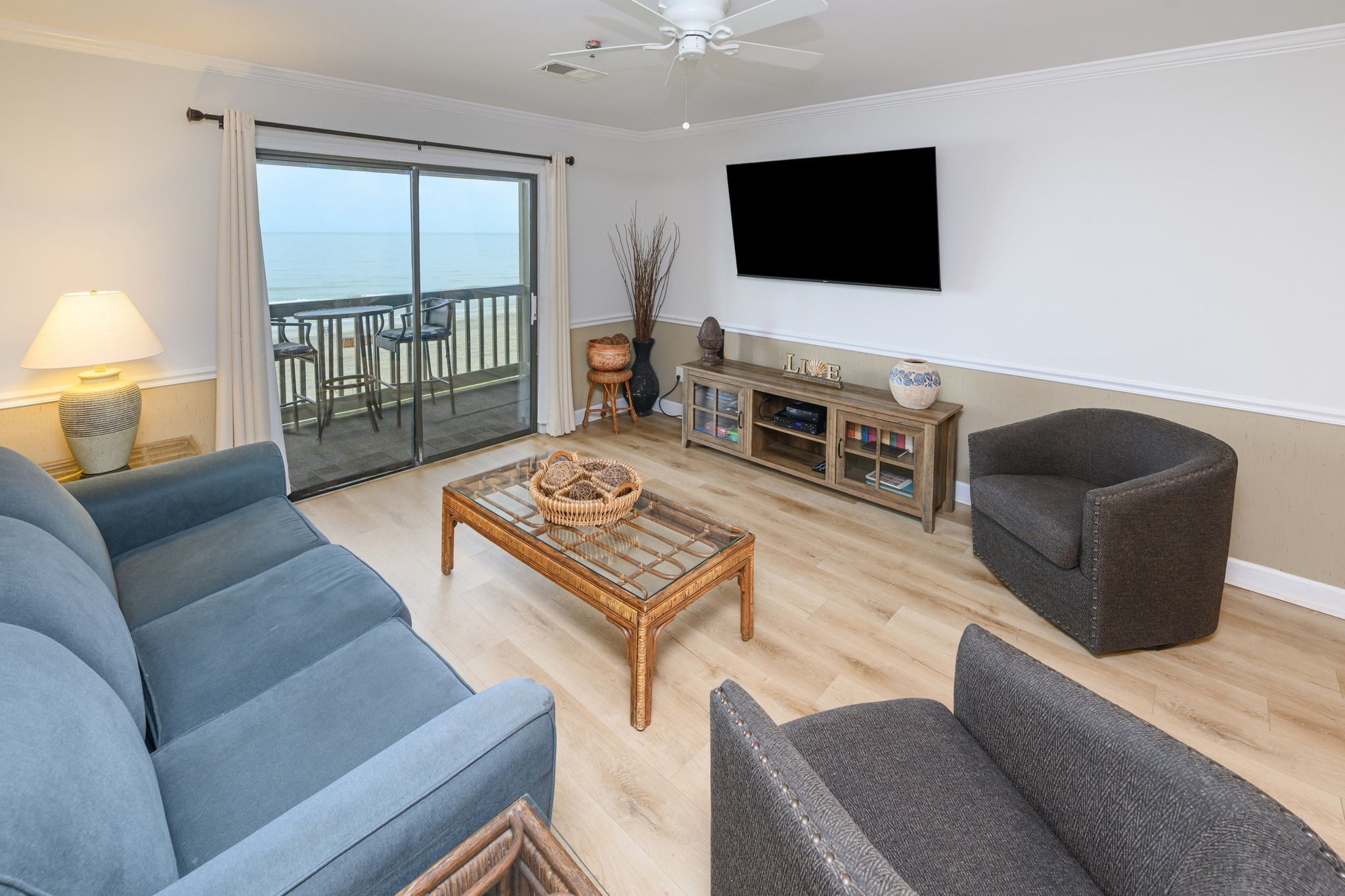 A living room with a sofa, armchair, coffee table, TV, and a view of the sea through a sliding door.