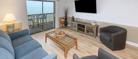 A living room with a sofa, armchair, coffee table, TV, and a view of the sea through a sliding door.
