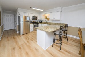 A modern kitchen with white cabinetry, a stainless steel refrigerator, a microwave, an oven, and a granite countertop island with bar stools. - 2BR Oceanfront Condo with Elevator and Shared Pool! Surfwatch II 302 (Surfside Beach)