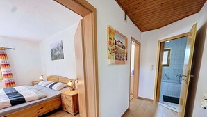Ein Schlafzimmer mit einem hölzernen Bett, einem Nachttisch und einem Beistelltisch. Eine Tür führt zu einem Badezimmer mit einer Dusche.