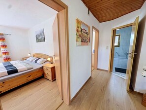Un dormitori amb llit de fusta, una taula de nit i un calaixera. Hi ha una porta que dona a un bany amb dutxa.