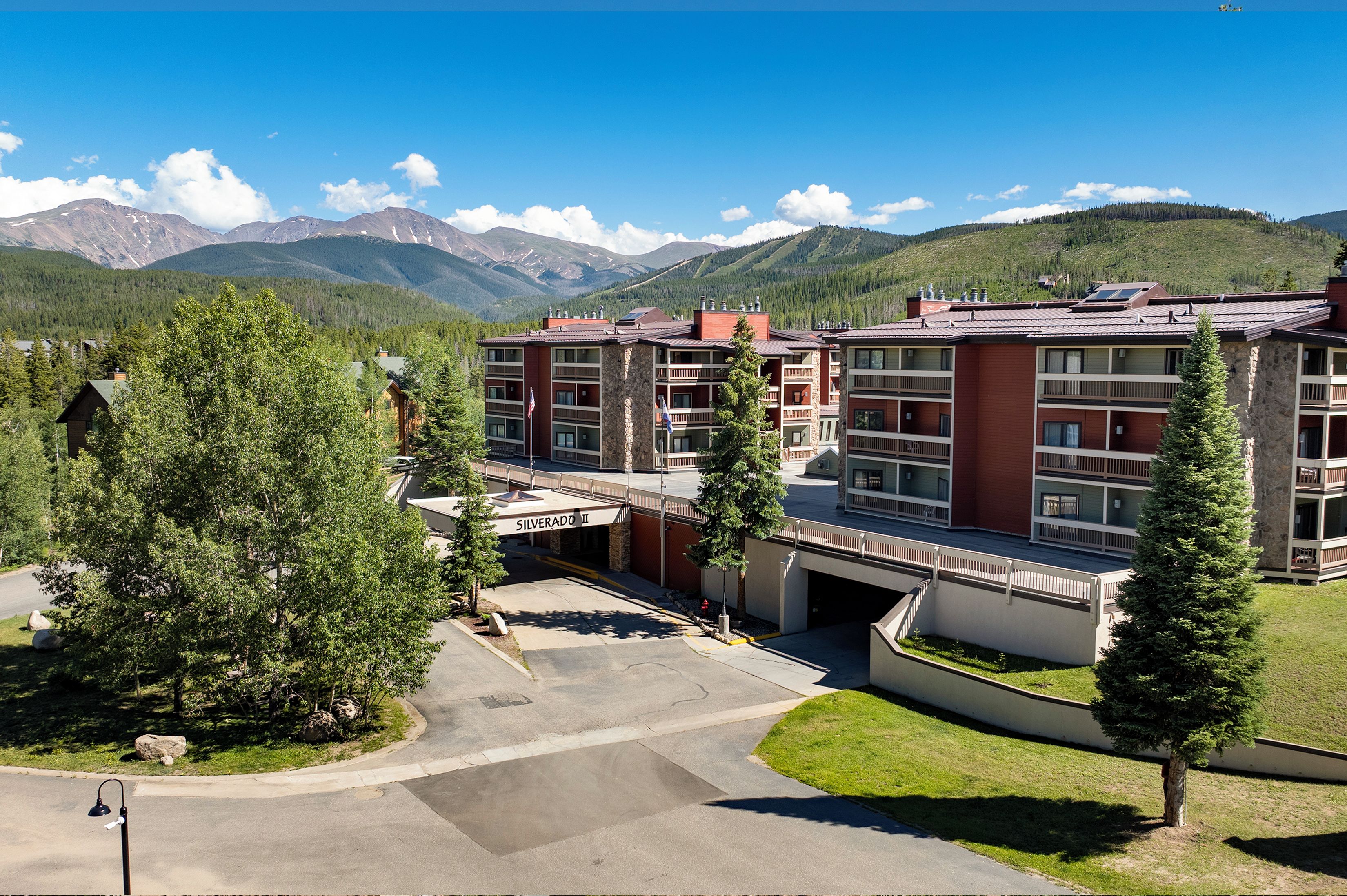 Silverado property nestled in a mountain valley, surrounded by evergreen forests and alpine peaks. The multi-level building features modern architecture with covered parking and a welcoming entrance.