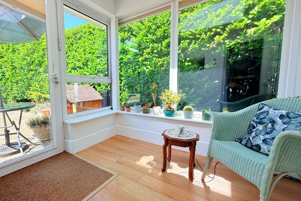 Una habitación iluminada por el sol con vistas a un follaje exuberante, una silla de mimbre, una pequeña mesa de madera y una estructura con techo de paja en el exterior.
