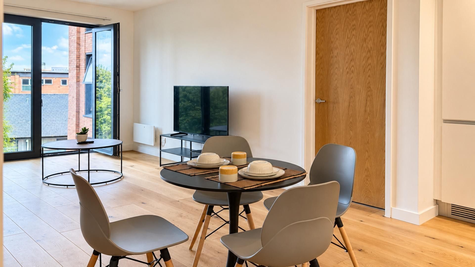 A modern living room with a round dining table, four chairs, a flat-screen TV, and a large window offering a view of buildings and greenery.