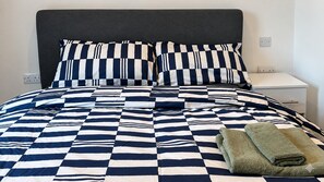 Ein Bett mit kariertem Überwurf und passenden Kissen, ein Nachttisch und ein gefaltetes Handtuch.