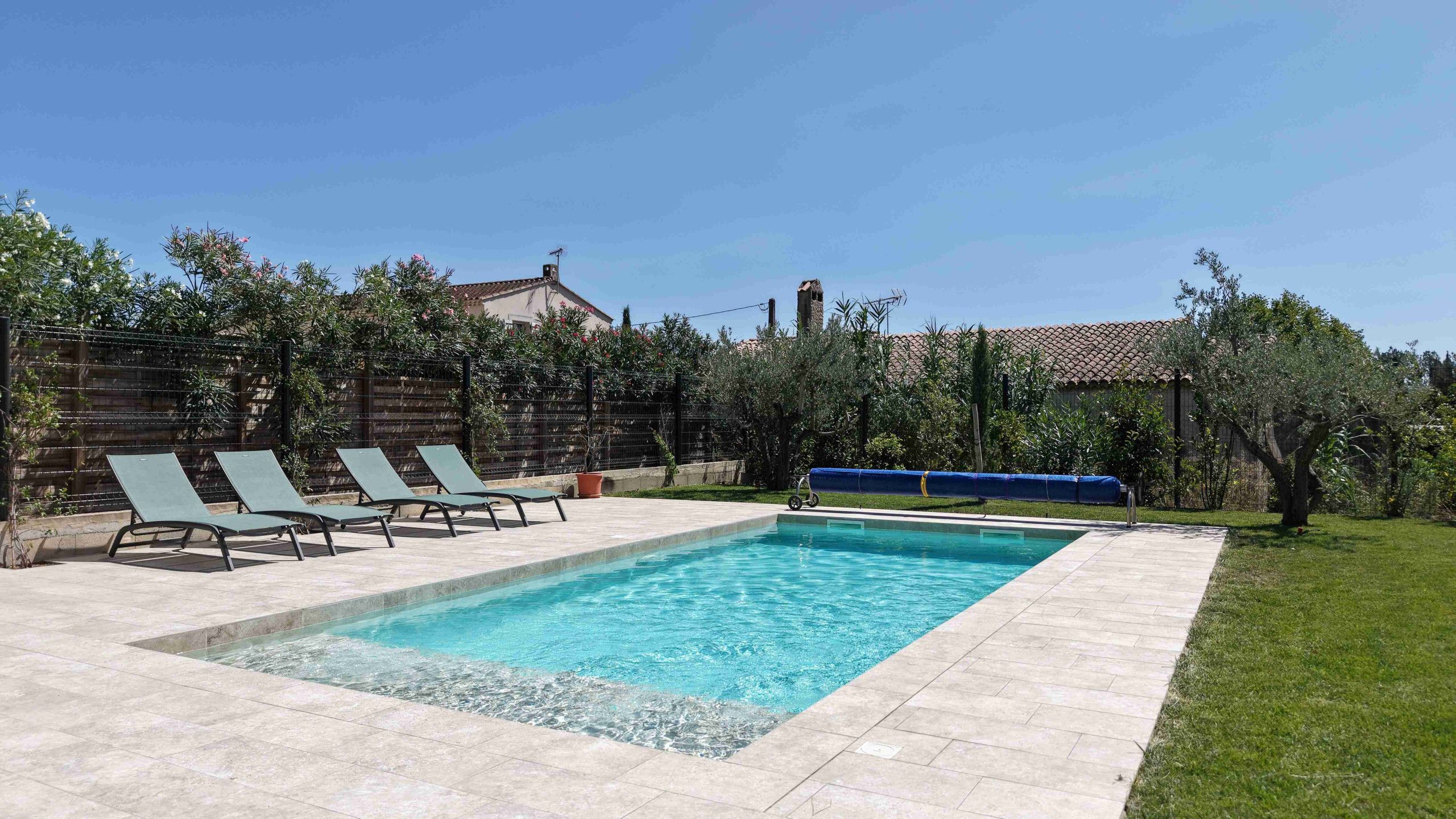Une piscine rectangulaire avec une terrasse carrelée, entourée d’une petite clôture et d’une végétation luxuriante.