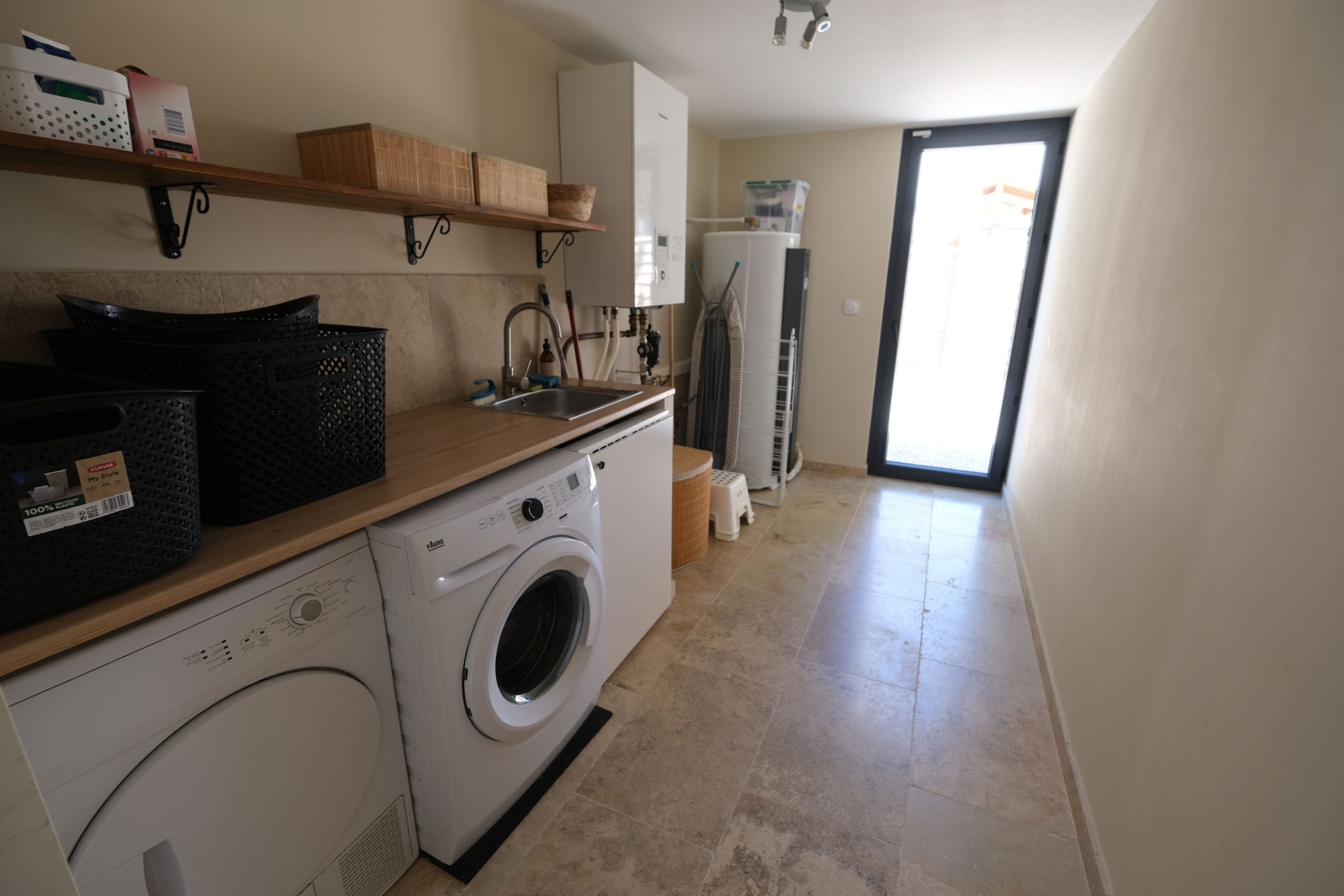 Zona de lavandería con lavadora, secadora y lavabo. Hay cestas y un estante con objetos.