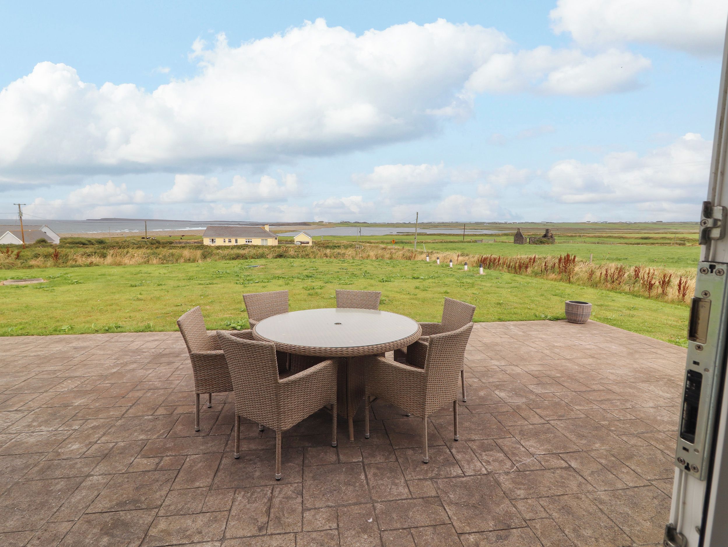 An outdoor patio with a round table and wicker chairs, overlooking a grassy field and a body of water.