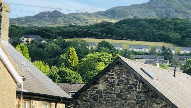 A stone building with a sloped roof, surrounded by lush greenery and houses in the background. - Tranquil Dolgellau Cottage Retreat (Dolgellau)