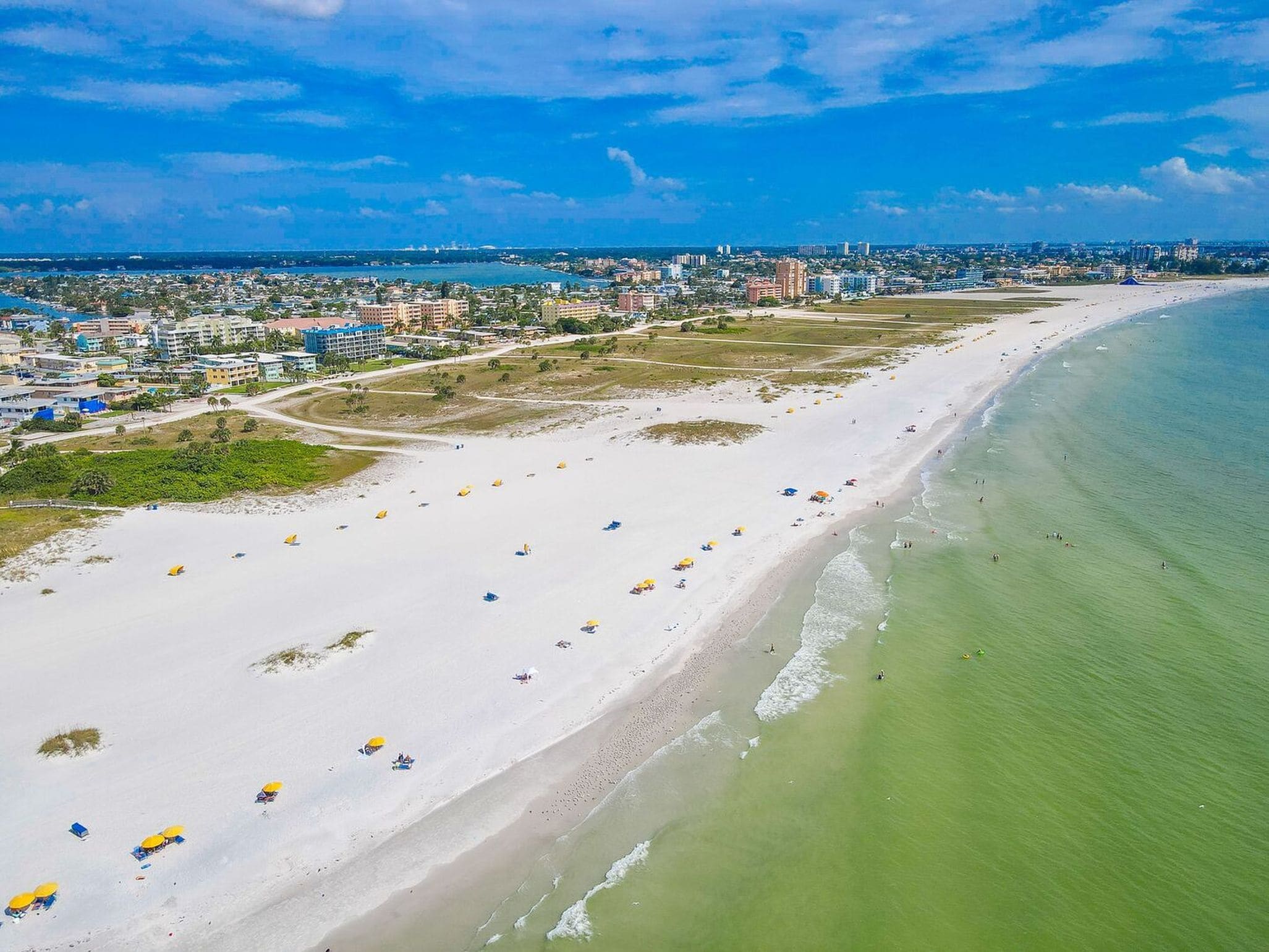A coastal cityscape with a wide sandy beach, a marina, and a dense urban area with buildings.