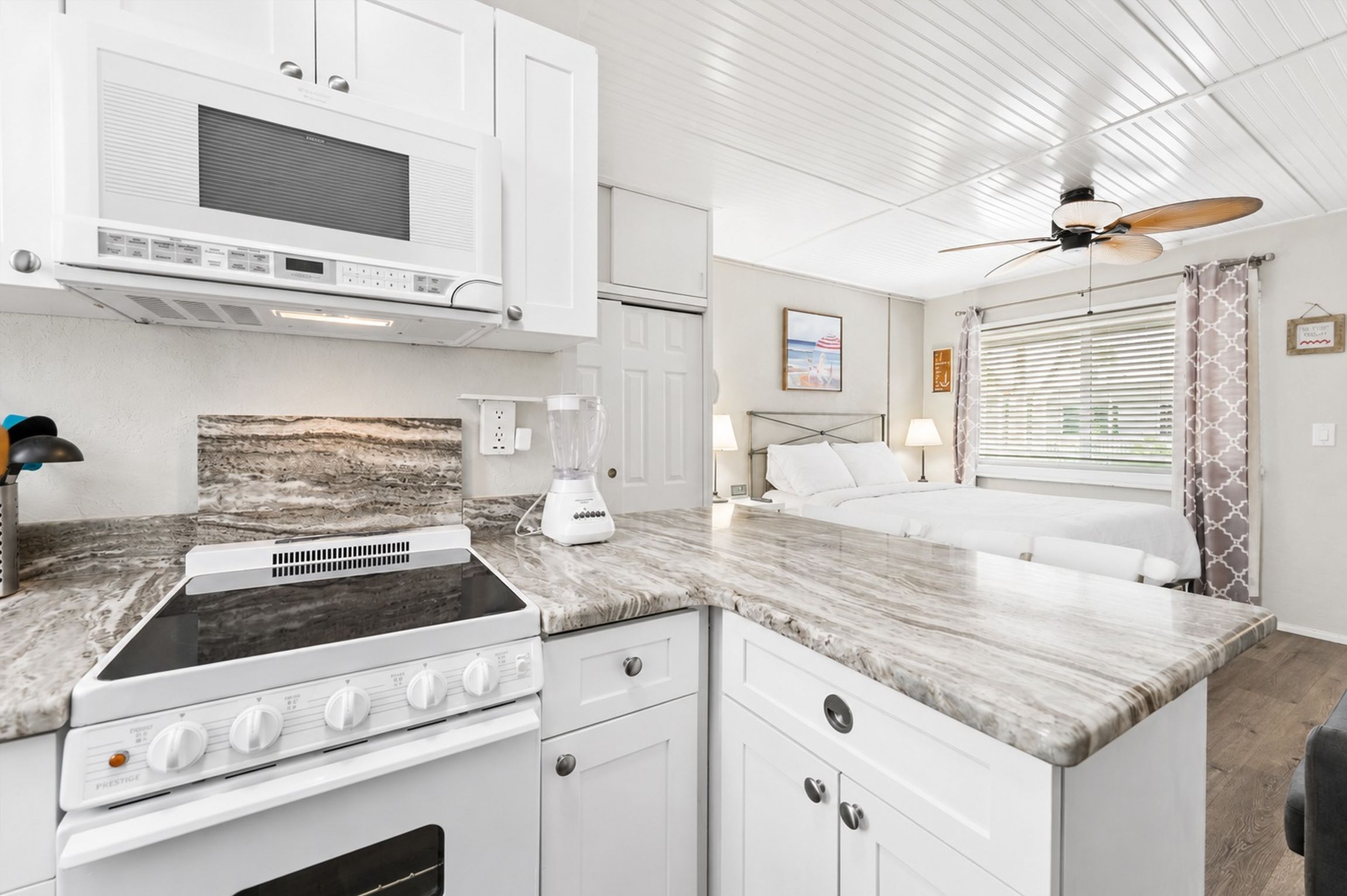 A compact kitchenette with a stove, microwave, and blender, a bed with a headboard, and a ceiling fan.