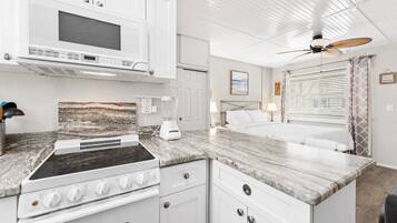A compact kitchenette with a stove, microwave, and blender, a bed with a headboard, and a ceiling fan.