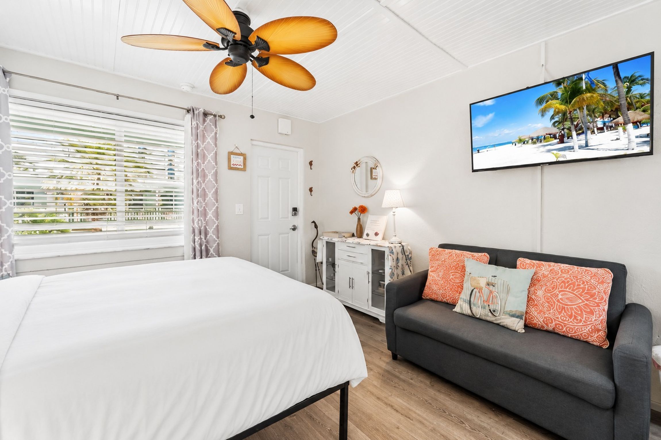 A hotel room with a bed, a sofa, a ceiling fan, a television, and a window with blinds.