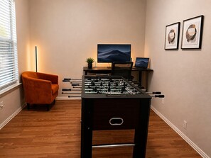 Ein Zimmer mit Kicker, einer Couch, einem Schreibtisch mit Computer und zwei gerahmten Bildern an der Wand.