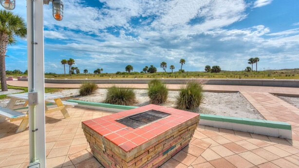 Un espacio al aire libre con pavimento, una jardinera de ladrillo, mesas de picnic y vista despejada a una calle y palmeras a lo lejos.