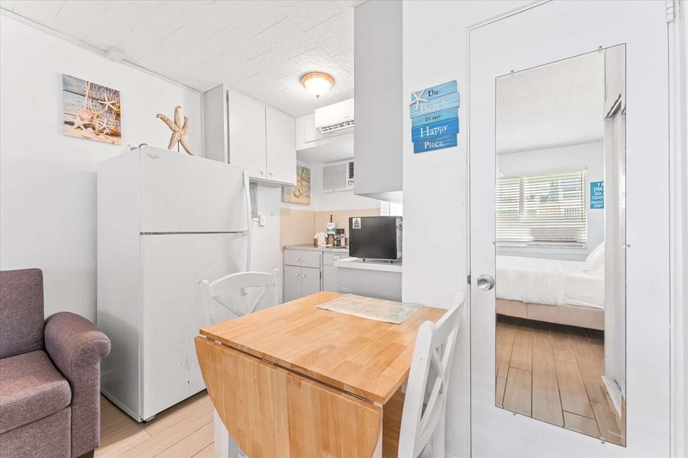 A compact kitchen with a refrigerator, a wooden table, and a chair. There is a bed with a white cover in the adjacent room.
