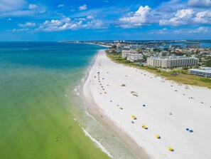 A beach with a long stretch of sand, a few umbrellas, and buildings in the background. - Tropic Terrace #56 | Pool View Studio | Resort Amenities (Treasure Island)