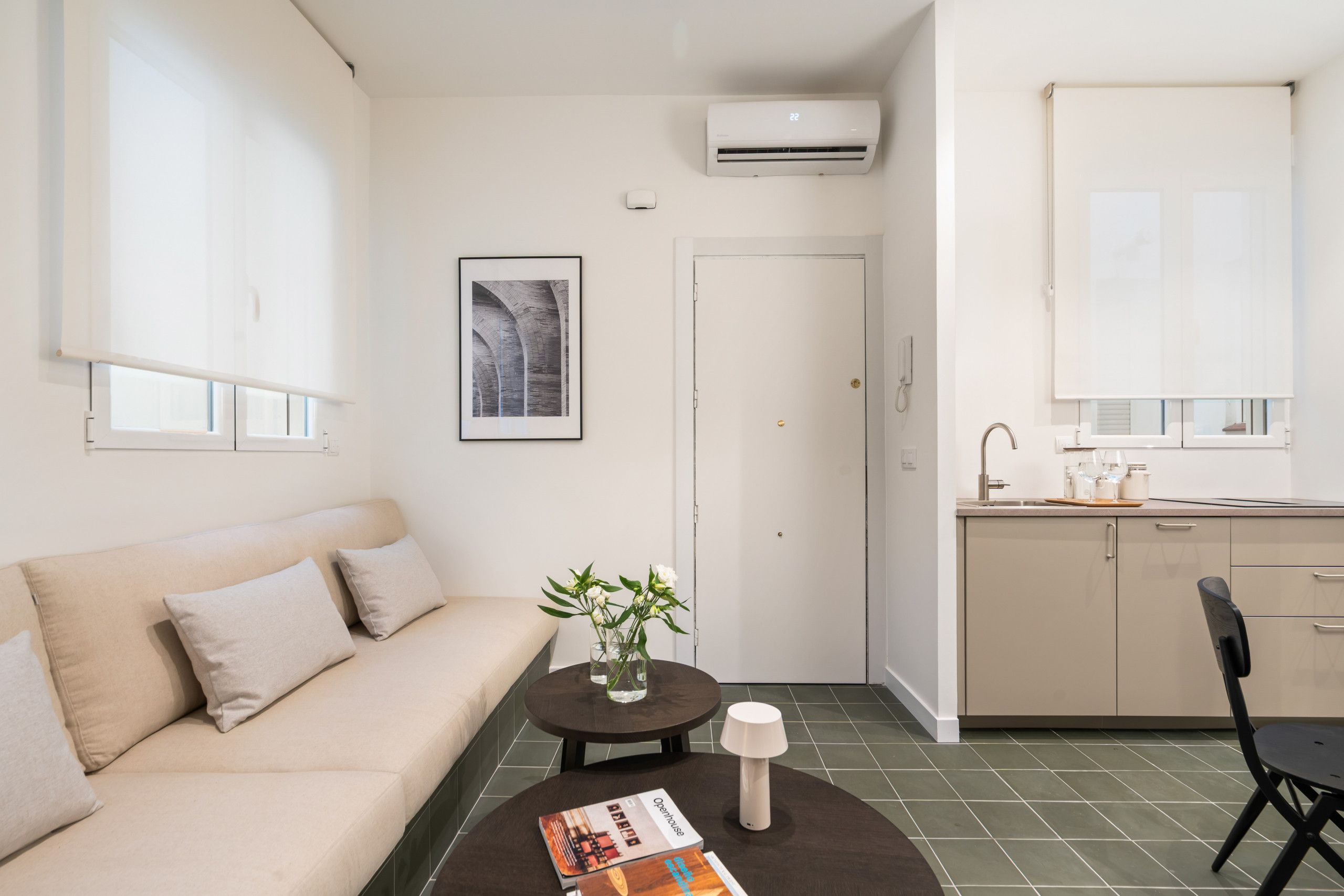 A modern, minimalist living space with a sofa, a small table with a vase of flowers, a framed picture on the wall, and a kitchen area with a window and a sink.