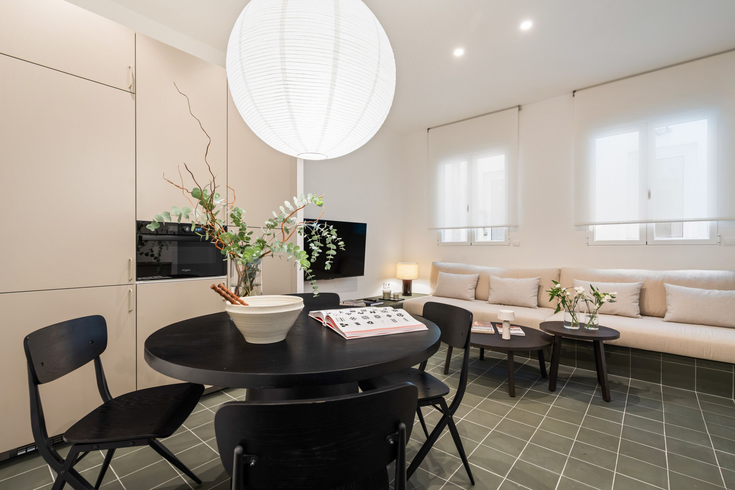 Een moderne woonkamer met een ronde eettafel, zwarte stoelen, een bank en een aan de muur bevestigde televisie.
