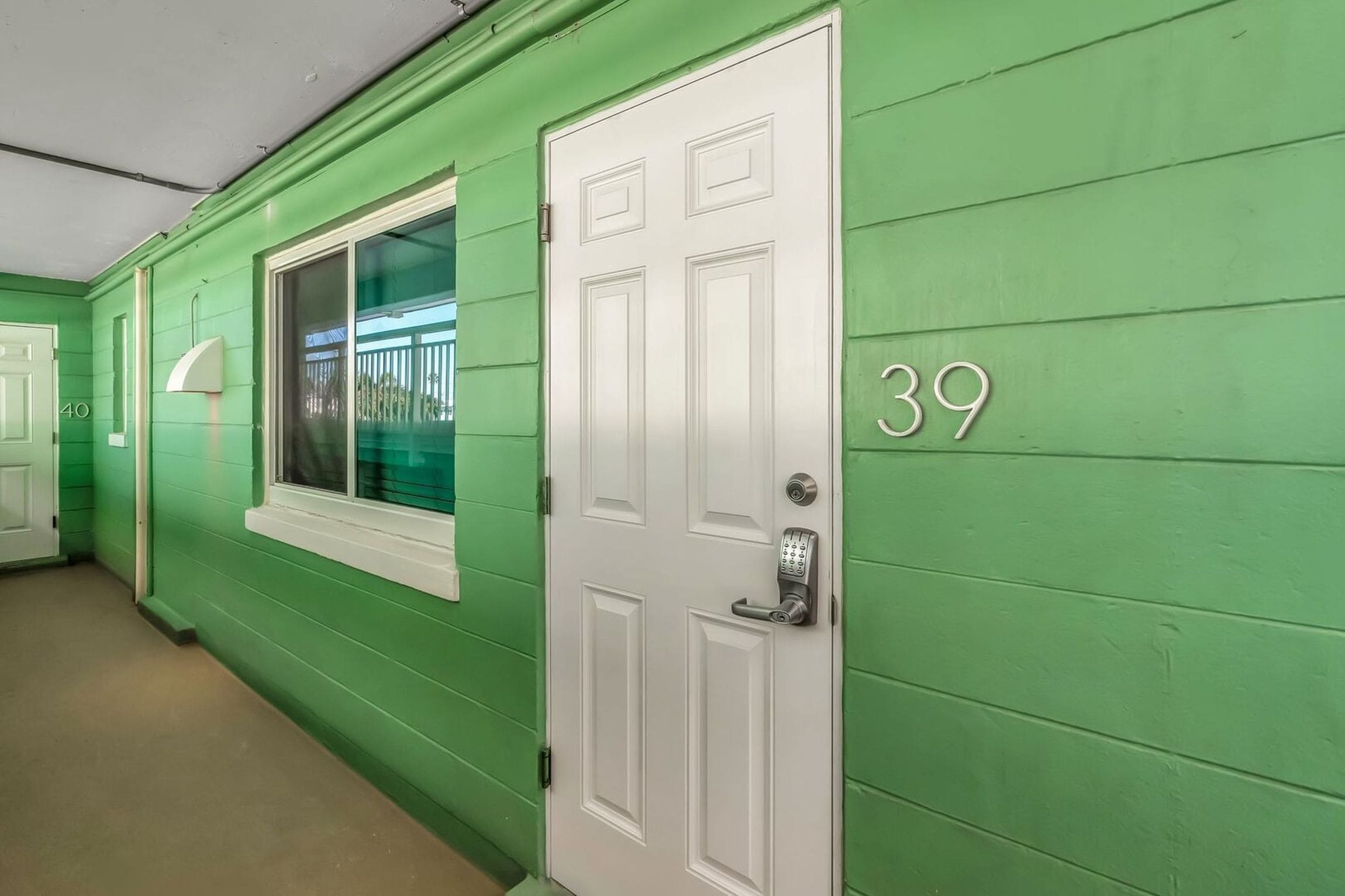 A hallway with green walls, a white door with a keypad lock, and a window with a view of a balcony.