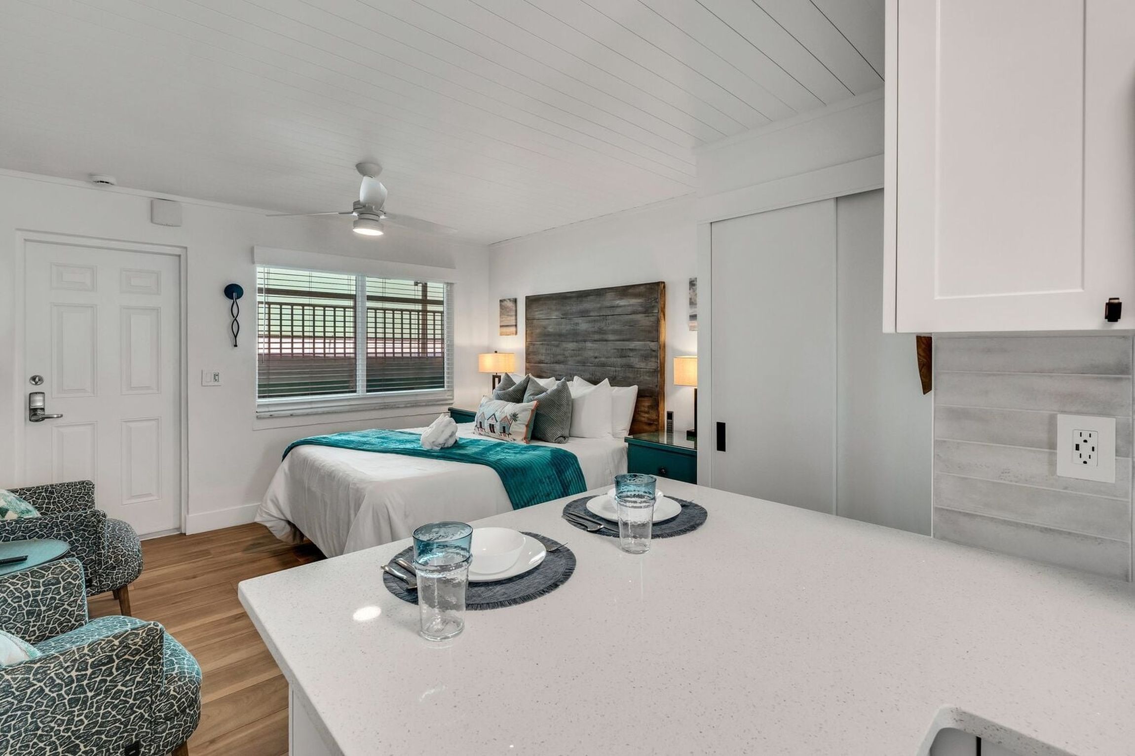 A modern hotel room with a bed, a dining table, and a window with blinds.