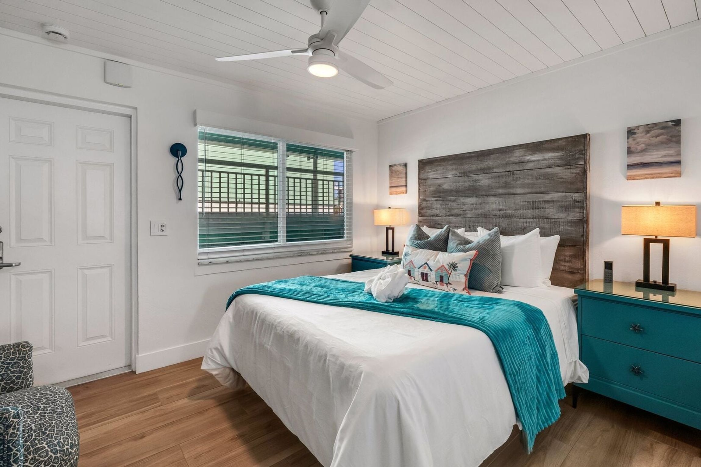A bedroom with a bed, two bedside tables, a ceiling fan, a window with blinds, and a wall-mounted artwork.