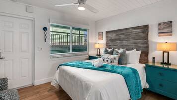 A bedroom with a bed, two bedside tables, a ceiling fan, a window with blinds, and a wall-mounted artwork.