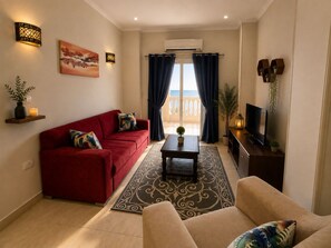 Een woonkamer met een rode bank, een beige fauteuil en een televisie op een houten dressoir. Er is een balkon met uitzicht, een bloempot en een decoratieve wandlamp.