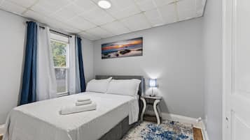 A bedroom with a bed, a nightstand, a lamp, a painting, and a window with curtains.