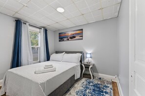A bedroom with a bed, a nightstand, a lamp, a painting, and a window with curtains. - Minutes To The Falls! Cozy Retreat Near State Park 3 Bedroom Apts (Niagara Falls)