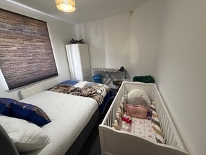 Quarto com cama, berço, radiador, janela com persianas, guarda-roupa e uma pequena mesa com cadeira.
