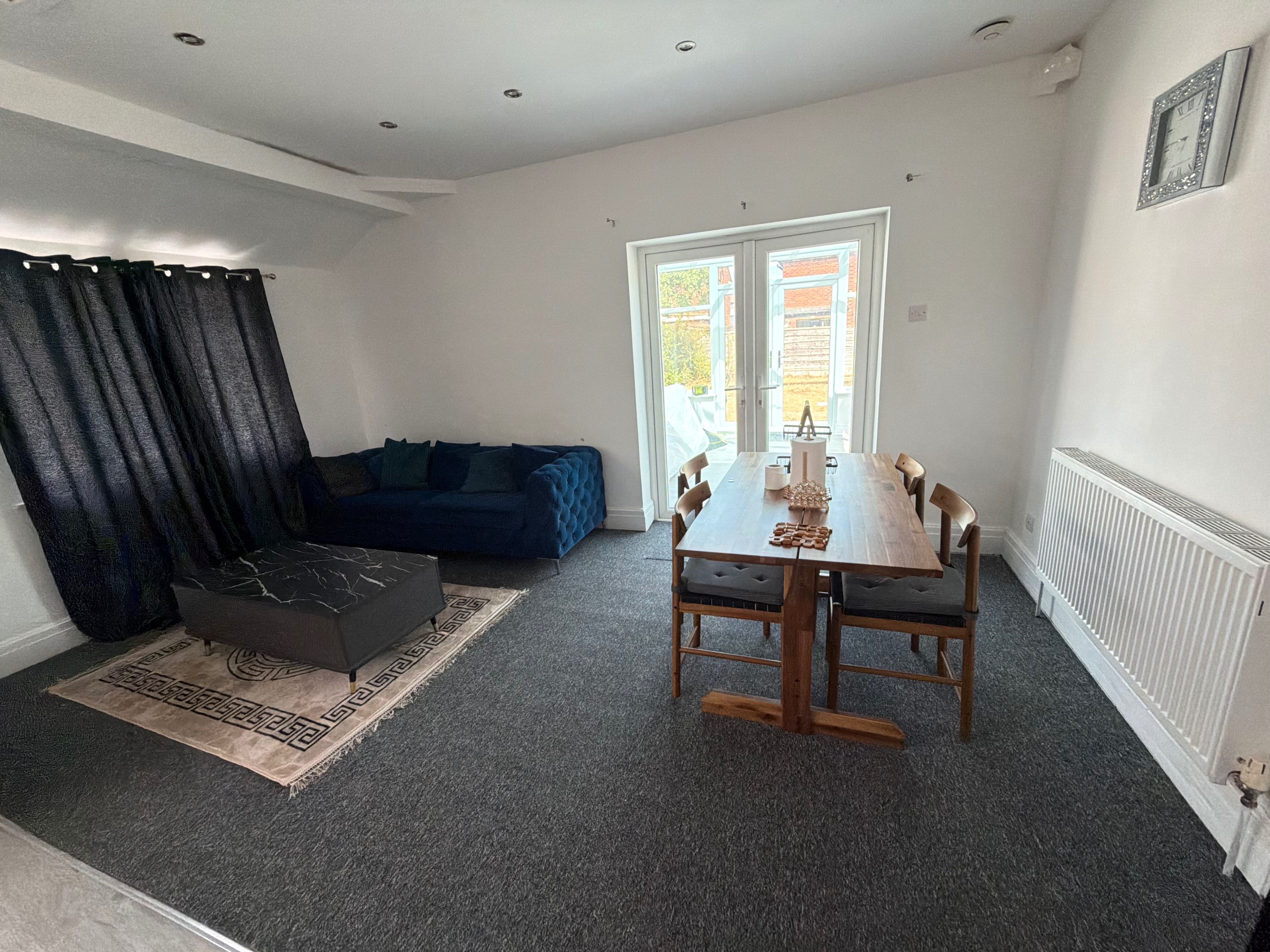 A modern living room with a dark blue sofa, a wooden dining table with chairs, a black ottoman, and a patterned rug.