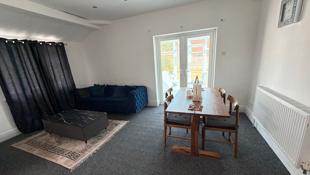 A modern living room with a dark blue sofa, a wooden dining table with chairs, a black ottoman, and a patterned rug. - House Sleeps 7 With Parking (Cheadle)