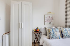 A bedroom with a bed, a bedside table with a fruit bowl, a white wardrobe, and a framed floral artwork on the wall. - Arsenal Corner Studio, 4 minutes to Kings Cross, with Ensuite & Self Check-In (London)