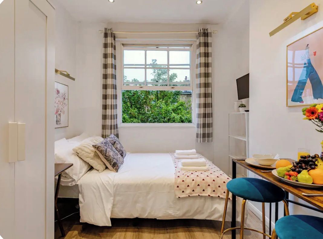 A compact bedroom with a bed, a small table with fruit, a TV, and a window with curtains.