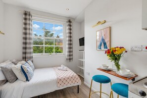A bedroom with a bed, a window with curtains, a TV, a painting, a vase with flowers, and a small table with a bowl and a cup. - Arsenal Corner Studio, 4 minutes to Kings Cross, with Ensuite & Self Check-In (London)