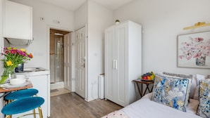 Ein kompaktes Hotelzimmer mit Küchenzeile, einem Bett mit blumigen Kissen, einem kleinen Tisch mit Obst und einem Gemälde an der Wand.