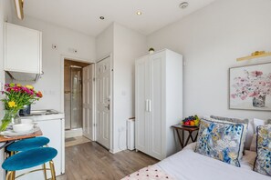 A compact hotel room with a kitchenette, a bed with floral-patterned pillows, a small table with fruit, and a painting on the wall. - Arsenal Corner Studio, 4 minutes to Kings Cross, with Ensuite & Self Check-In (London)